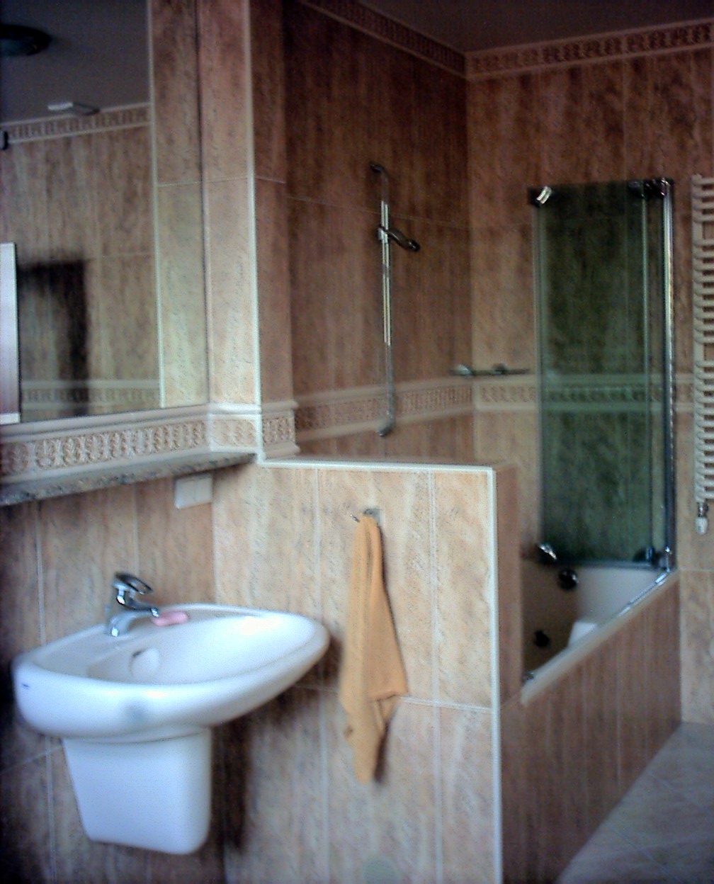 Ukończona łazienka z beżowymi płytkami, umywalką na postumencie, wanną z parawanem i lustrem. Na ścianie wisi pomarańczowy ręcznik.