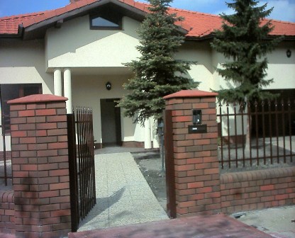 Dom jednorodzinny z beżową elewacją, czerwoną dachówką i ogrodzeniem z ceglanymi słupkami oraz metalową bramką. Widoczne dwie choinki przed domem.