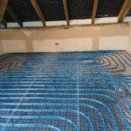 Niebieskie rurki ogrzewania podłogowego ułożone spiralnie na siatce, widoczne fragmenty ścian i drewnianej konstrukcji dachu.