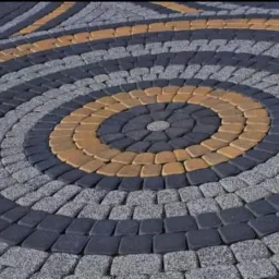 Okrągły wzór z kostki brukowej w odcieniach szarości, grafitu i beżu, tworzący dekoracyjny element nawierzchni.