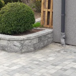 Kaskadowy kwietnik z szarej kostki, otaczający kulisty krzew, na tle szarej kostki brukowej i ściany budynku z rynną.