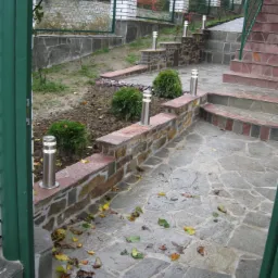 Kamienny taras z nieregularnych płyt, oświetlony niskimi lampami i ozdobiony niskimi krzewami, prowadzący do schodów z bordowymi akcentami, otoczony ogrodzeniem z białej cegły.