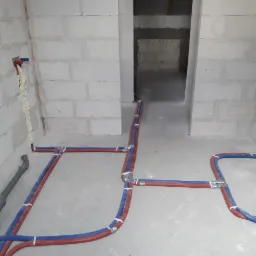 Wnętrze w trakcie prac hydraulicznych: rury grzewcze PEX w kolorze czerwono-niebieskim, łączniki, pianka montażowa uszczelniająca przejścia przez ścianę z betonu komórkowego.