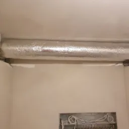 Ocieplony, srebrną folią aluminiową kanał wentylacyjny pod sufitem w trakcie montażu, widoczne niedokończone ściany i wystające kable.