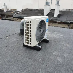 Kompaktowa jednostka zewnętrzna klimatyzacji marki Midea, umieszczona na dachu pokrytym papą, z widocznymi czarnymi podkładkami antywibracyjnymi i szarym przewodem odprowadzającym.