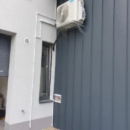 Zewnętrzna jednostka klimatyzatora Midea zamontowana na metalowej elewacji budynku, widoczne rury instalacyjne prowadzone wzdłuż ściany i mały zawór z kranikiem.