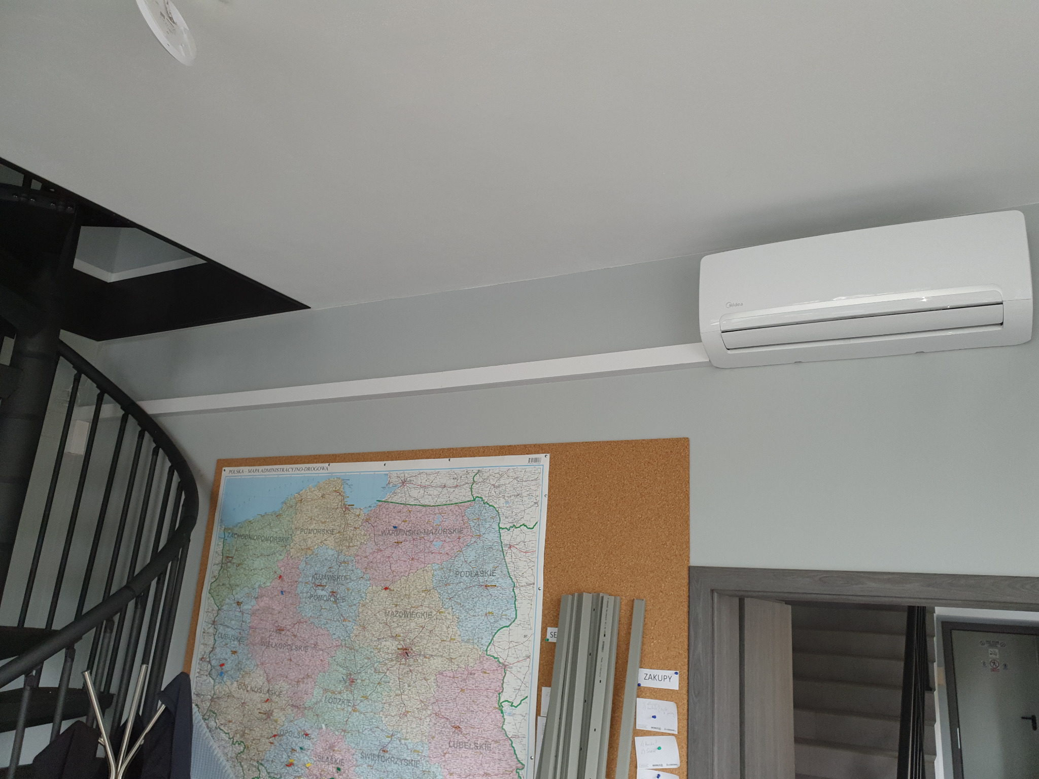 Biały klimatyzator Midea zamontowany na szarej ścianie, poniżej mapa Polski na korkowej tablicy i fragment spiralnych schodów.