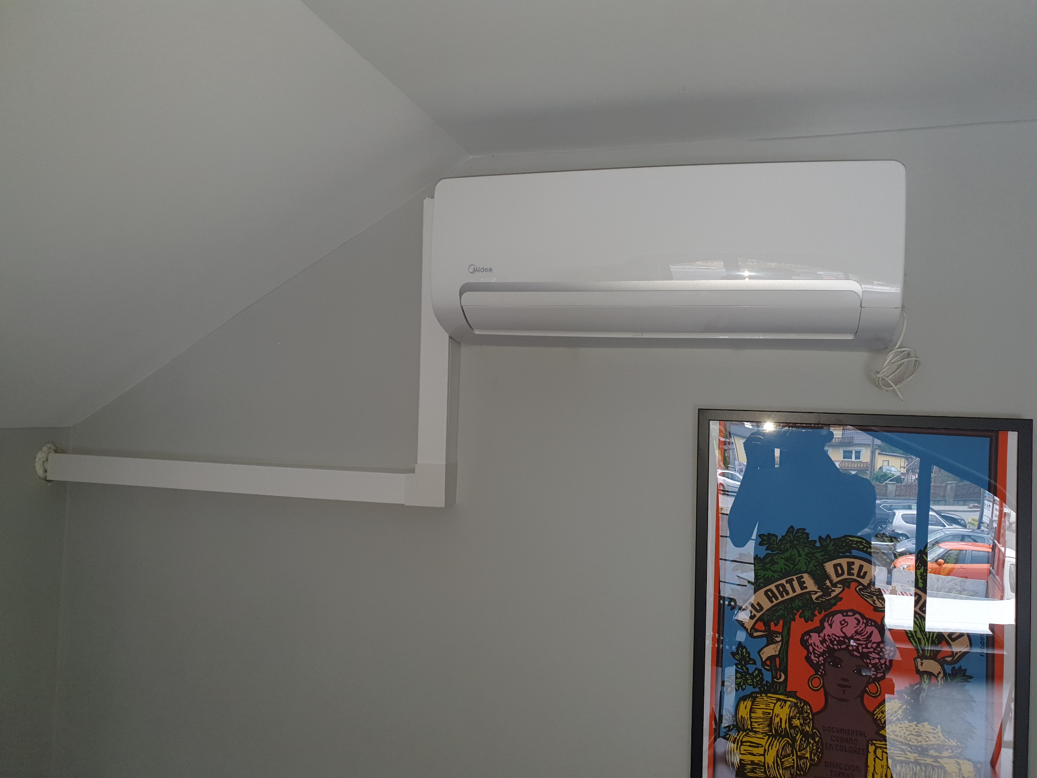 Biały klimatyzator marki Midea zamontowany na szarej ścianie w pomieszczeniu ze skośnym sufitem, obok widoczna rama obrazu z refleksami otoczenia.