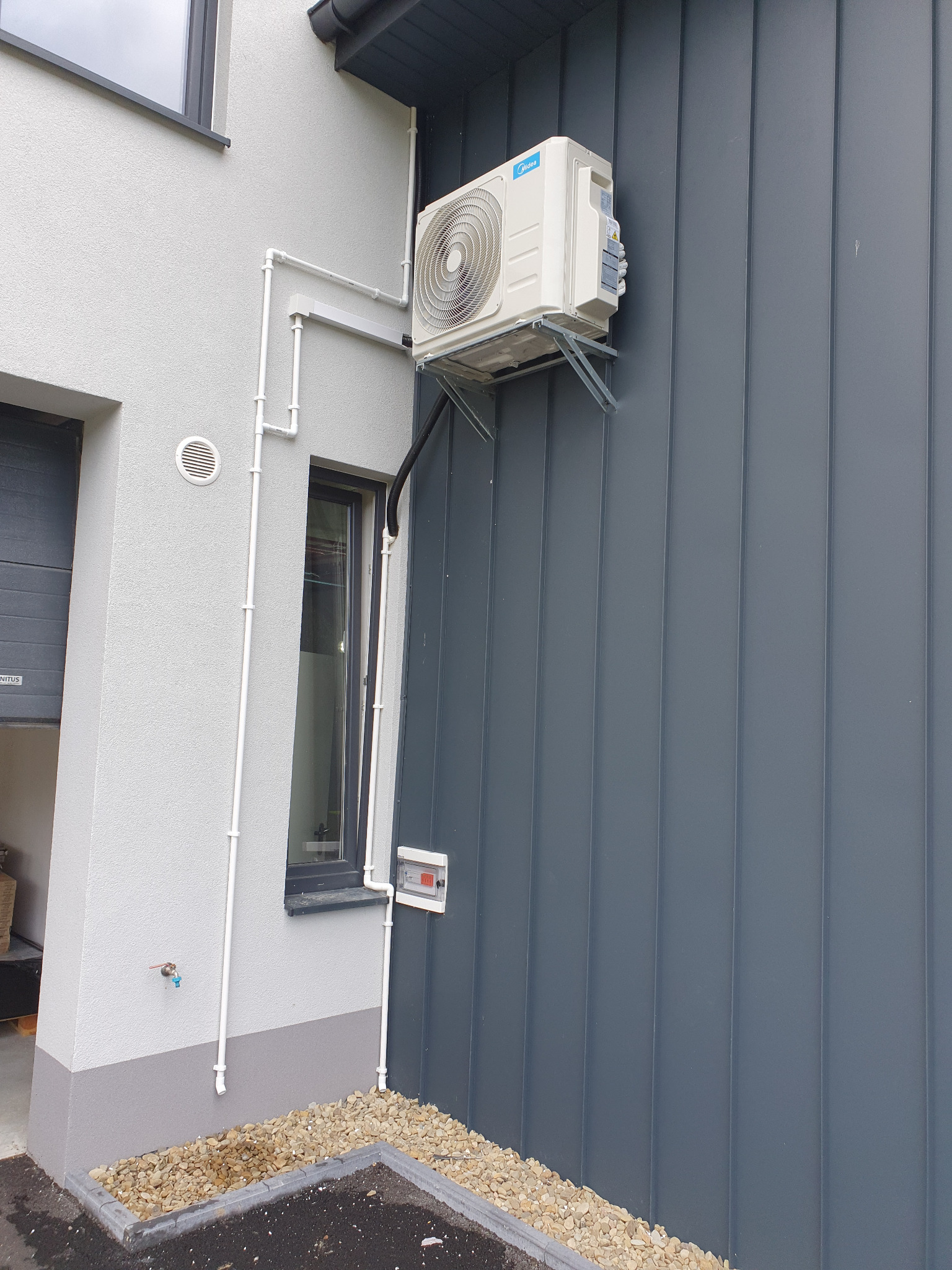 Zewnętrzna jednostka klimatyzatora Midea zamontowana na metalowej elewacji budynku, widoczne rury instalacyjne prowadzone wzdłuż ściany i mały zawór z kranikiem.