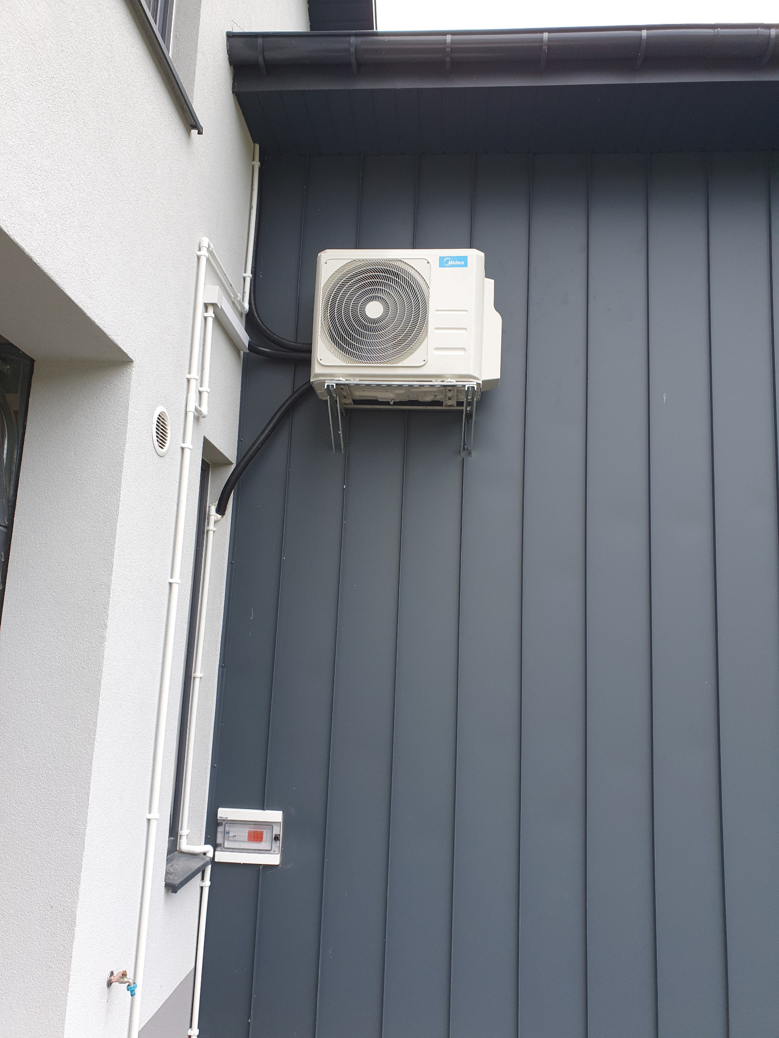 Biała jednostka zewnętrzna klimatyzacji Midea zamontowana na szarej elewacji z blachy trapezowej, widoczne rury instalacyjne prowadzone wzdłuż ściany budynku.