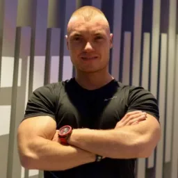 Uśmiechnięty mężczyzna o krótkich włosach w czarnej koszulce, z założonymi rękami i czerwonym zegarkiem na tle ściany z pionowymi panelami.