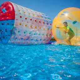 Dwie duże, przezroczyste kule do chodzenia po wodzie: jedna wielokolorowa, druga pomarańczowa z chłopcem w środku, na basenie w słoneczny dzień.