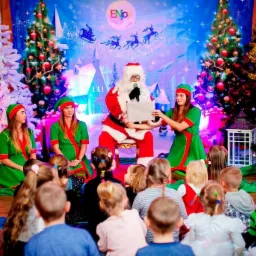 Scena z Mikołajem czytającym list w otoczeniu dwóch pomocnic i grupy dzieci, bożonarodzeniowa dekoracja sceniczna z choinkami w tle.