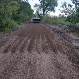 Żółta spycharka wyrównuje żwirową drogę gruntową, widoczne ślady po przejeździe maszyny, w tle drzewa.