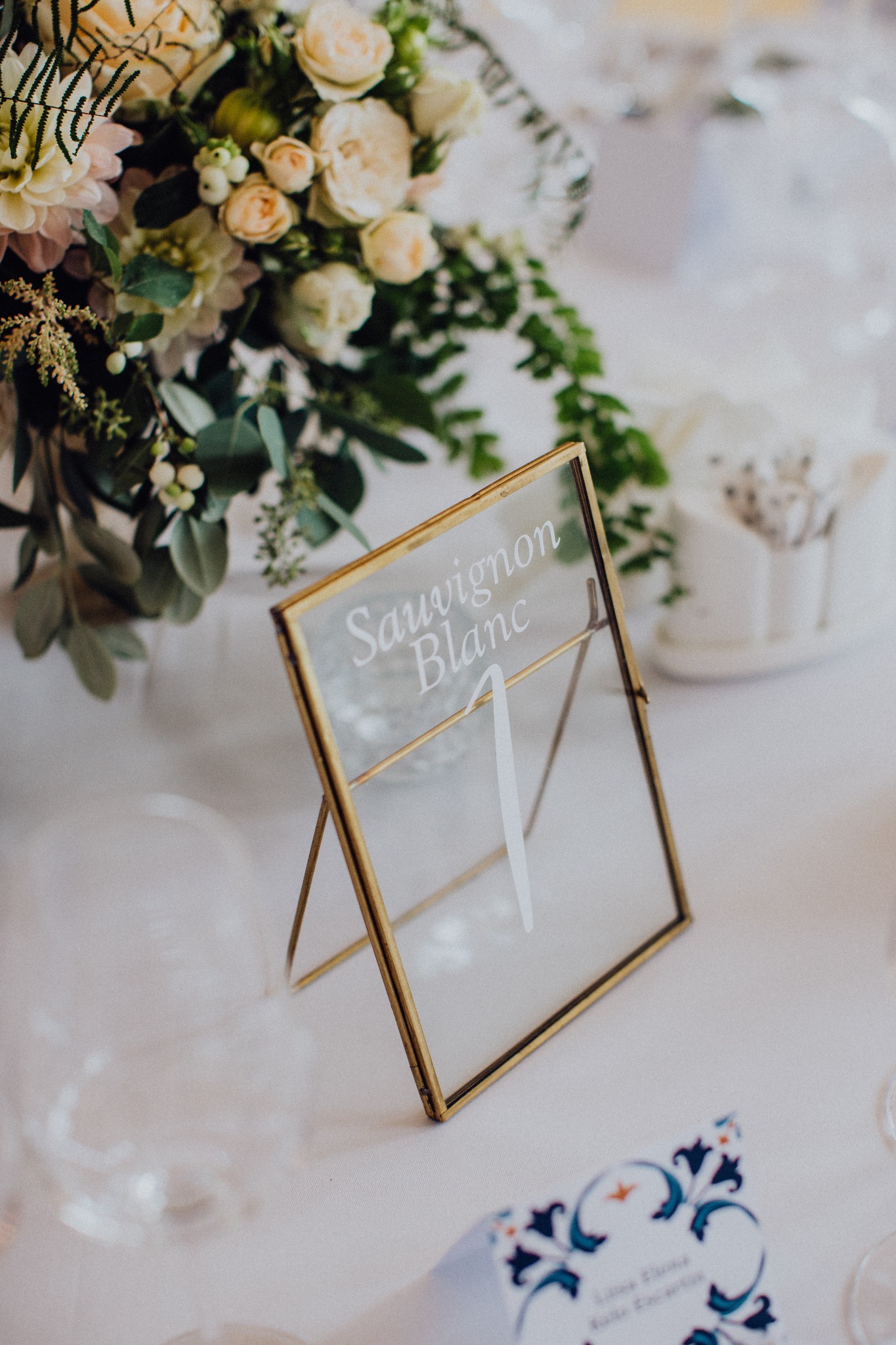 Dekoracja stołu weselnego: złota ramka z napisem 'Sauvignon Blanc' i numerem '1', bukiet kwiatów w tle, winietka z niebieskim wzorem.
