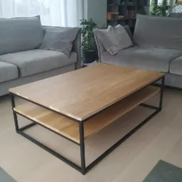 Stolik kawowy z blatem z jasnego drewna i czarną metalową ramą, z dwiema półkami, ustawiony w salonie z szarymi sofami i rośliną doniczkową w tle.