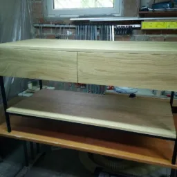 Niska komoda z dwoma szufladami z jasnego drewna na czarnych, metalowych nogach, z dodatkową półką, ustawiona w warsztacie.