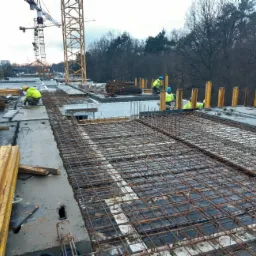 Zbrojenie stropu na budowie z pracownikami w odblaskowych ubraniach i dźwigami w tle.