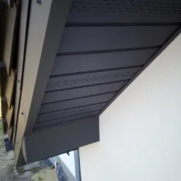 Szczegół zadaszenia budynku z widocznymi panelami z perforacją wentylacyjną, mocowanymi na śruby, przylegający do jasnej ściany z widoczną krawędzią okna.