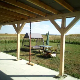 Drewniana konstrukcja zadaszenia tarasu, widok na pole i huśtawkę ogrodową w słoneczny dzień.