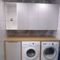 Białe, lakierowane szafki wiszące nad blatem drewnianym z pralką i suszarką. Jedna szafka z przeszklonym frontem. Szare płytki na ścianie w tle.