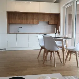 Jasna kuchnia z białymi frontami i drewnianymi szafkami wiszącymi, oświetlona czarnymi reflektorami, z okrągłym stołem i krzesłami w stylu skandynawskim, widok z salonu na otwartą przestrzeń.