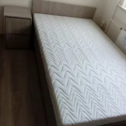 Łóżko z jasnego drewna z materacem o wzorze falistym, obok szafka nocna z dwoma szufladami, widoczne drewniane panele podłogowe.