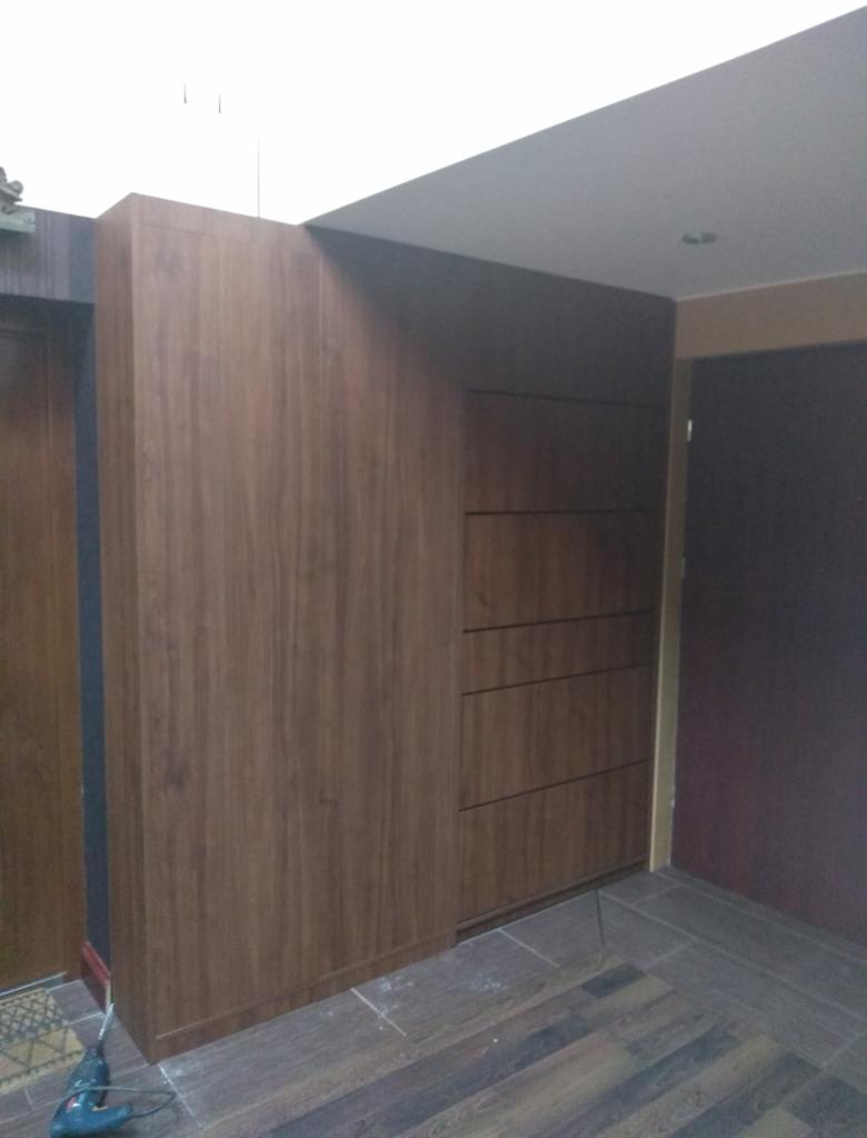 Wnętrze z zabudowaną szafą z drewnopodobnymi frontami, widoczna szuflada z poziomymi uchwytami, w tle drzwi i podłoga z płytek imitujących drewno, w rogu wiertarka.