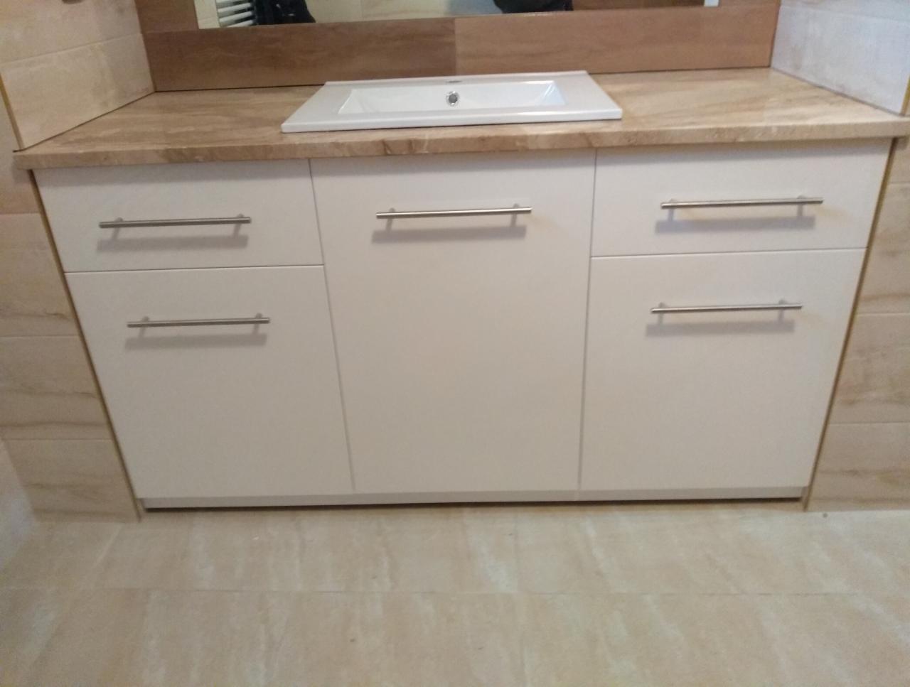 Biała szafka łazienkowa pod umywalką z beżowym blatem imitującym marmur i podłużnymi, metalowymi uchwytami, na tle beżowych płytek.