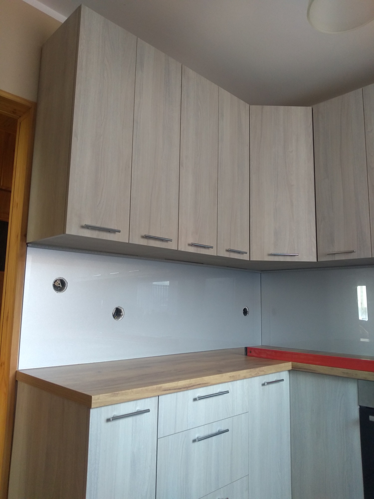 Jasne, drewnopodobne szafki kuchenne podwieszane z szarym blatem i białym panelem szklanym, widoczne otwory na gniazdka elektryczne, poziomica na blacie.