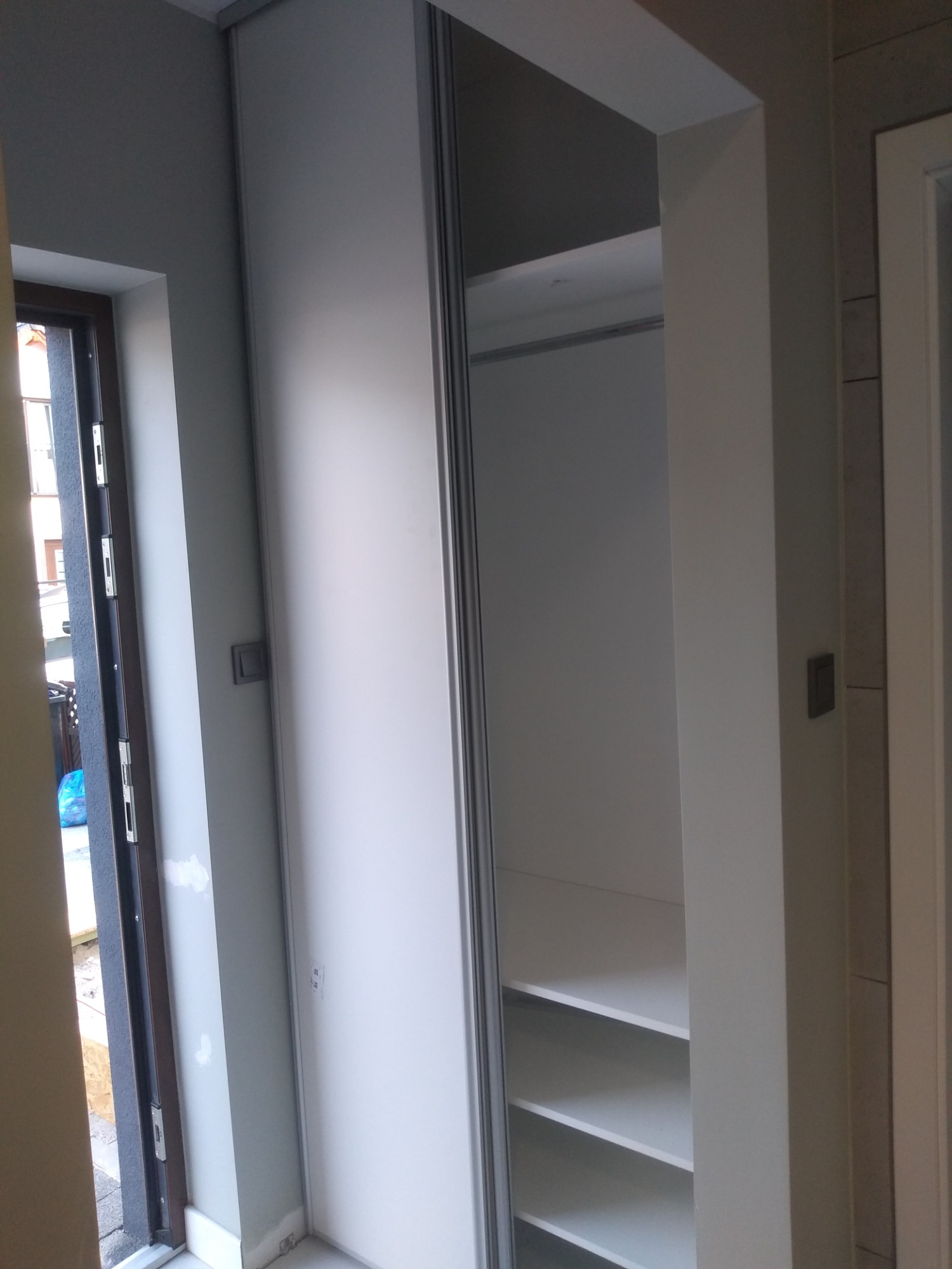 Biała szafa wnękowa z przesuwnymi drzwiami, widoczne półki wewnątrz, fragment okna i drzwi w tle.