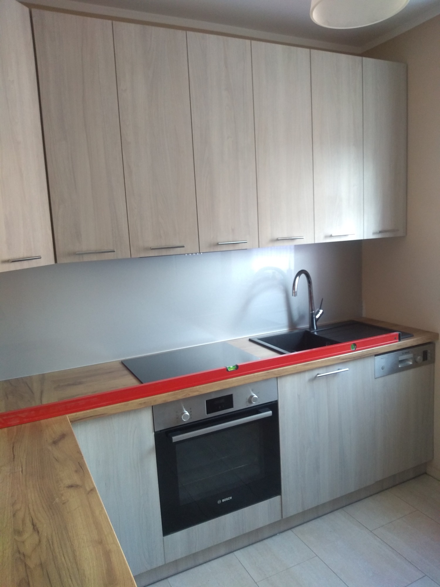 Jasna zabudowa kuchenna z drewnianymi frontami i szarym blatem, poziomowana z użyciem poziomicy w kolorze czerwonym, widoczny zlew, płyta indukcyjna i piekarnik Bosch.