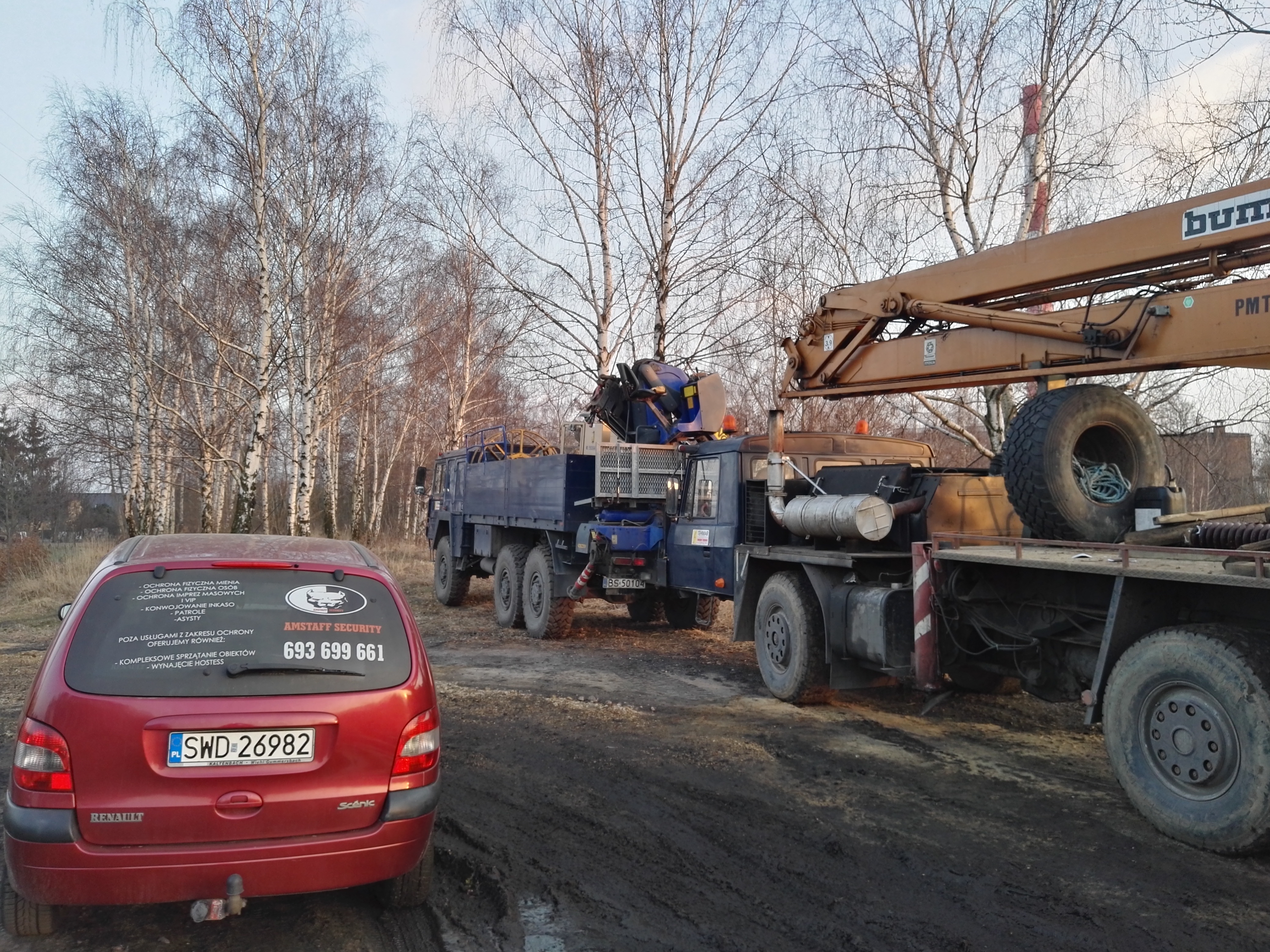 Czerwony samochód Renault Scenic z naklejką Agencji Ochrony Amstaff Security na tylnej szybie, zaparkowany na błotnistej drodze, w tle ciężarówka z dźwigiem i brzozowy zagajnik.