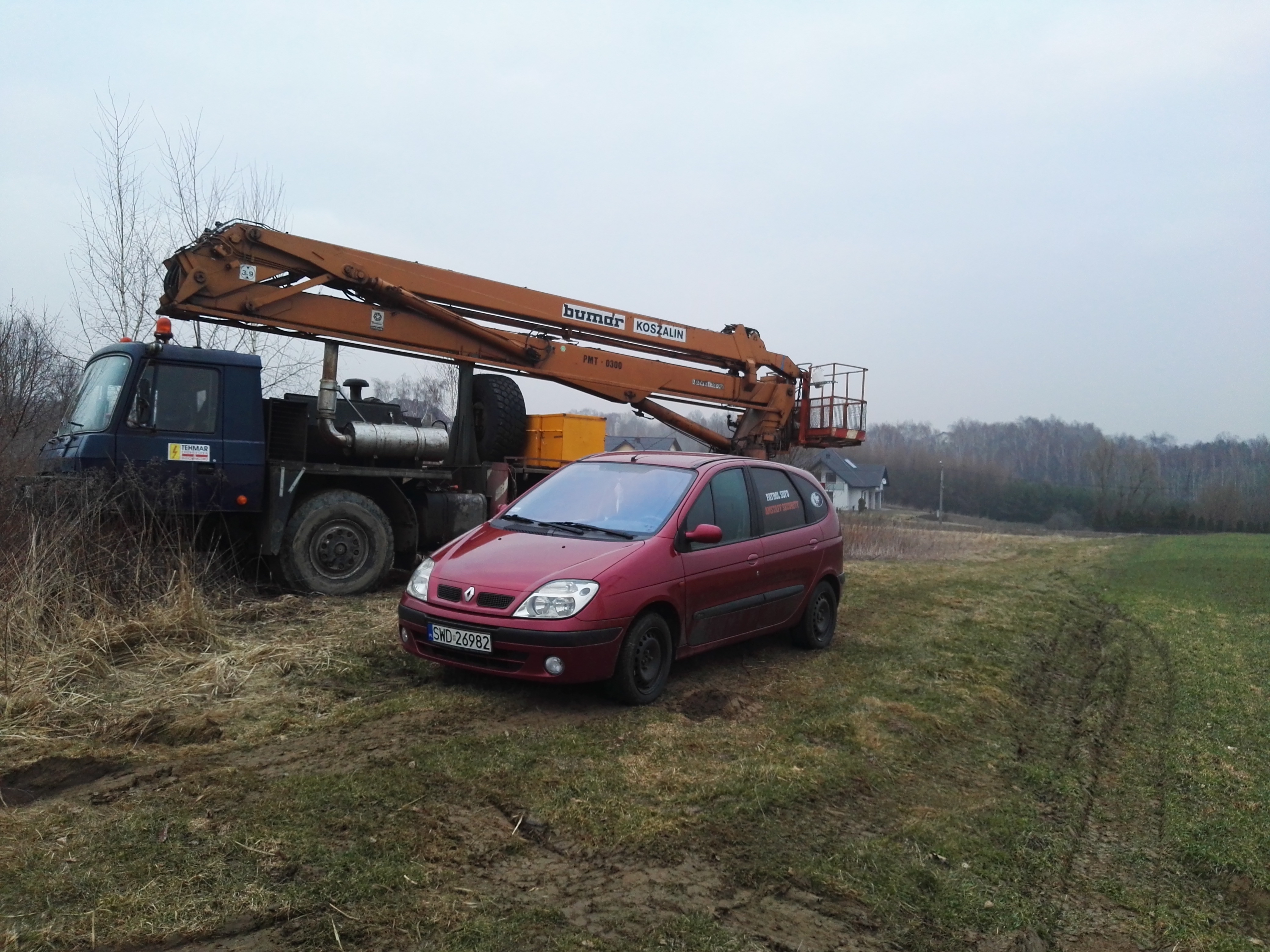 Czerwony samochód osobowy marki Renault z naklejką 'PATROL SUFO AMSTAFF SECURITY' na szybie, zaparkowany obok dźwigu samochodowego Bumar Koszalin na terenie podmokłym.