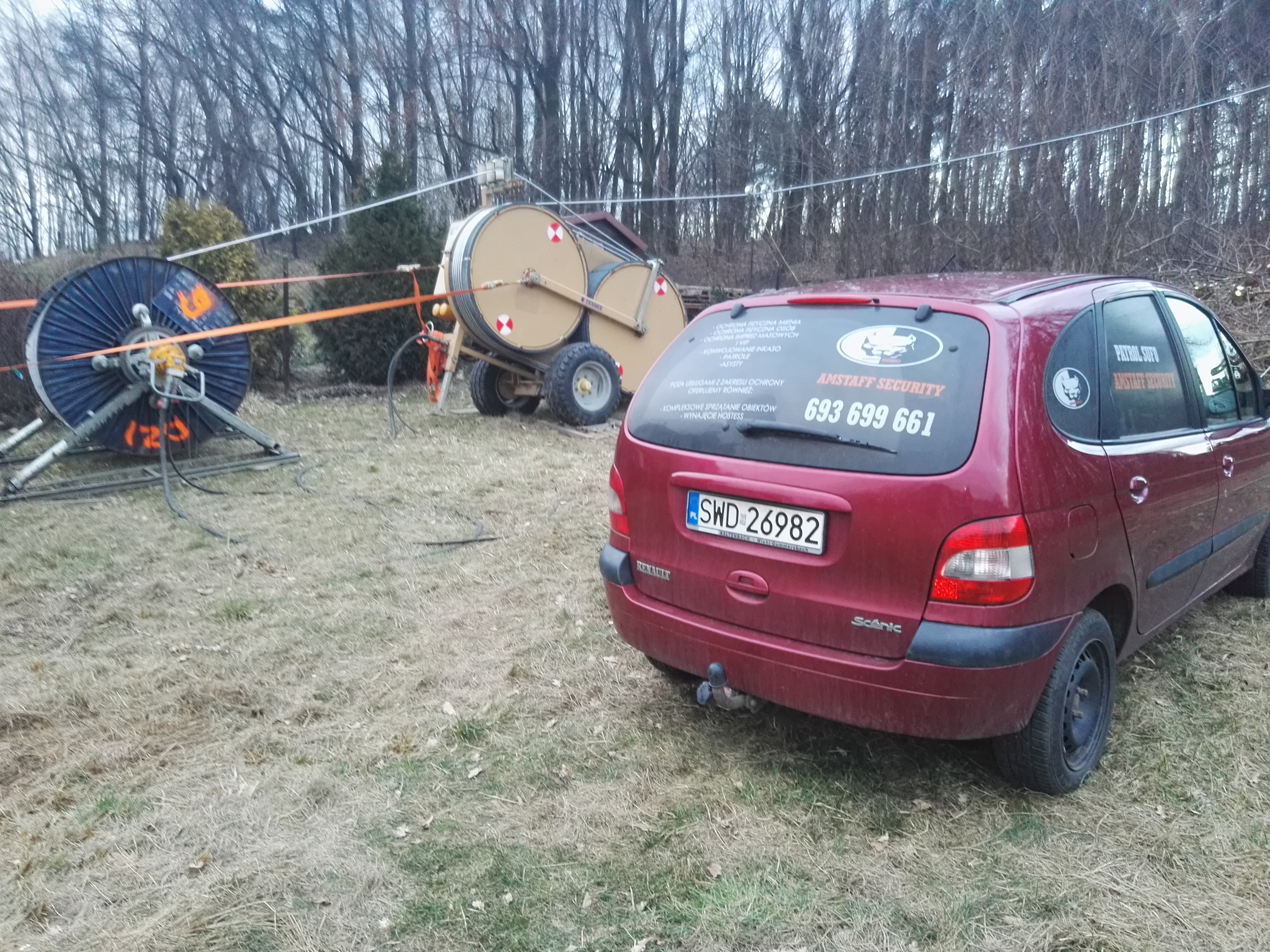 Czerwony samochód Renault Scenic z naklejkami firmy ochroniarskiej Amstaff Security na tylnej szybie, zaparkowany na trawiastym terenie z widocznymi dwoma bębnami kablowymi w tle.