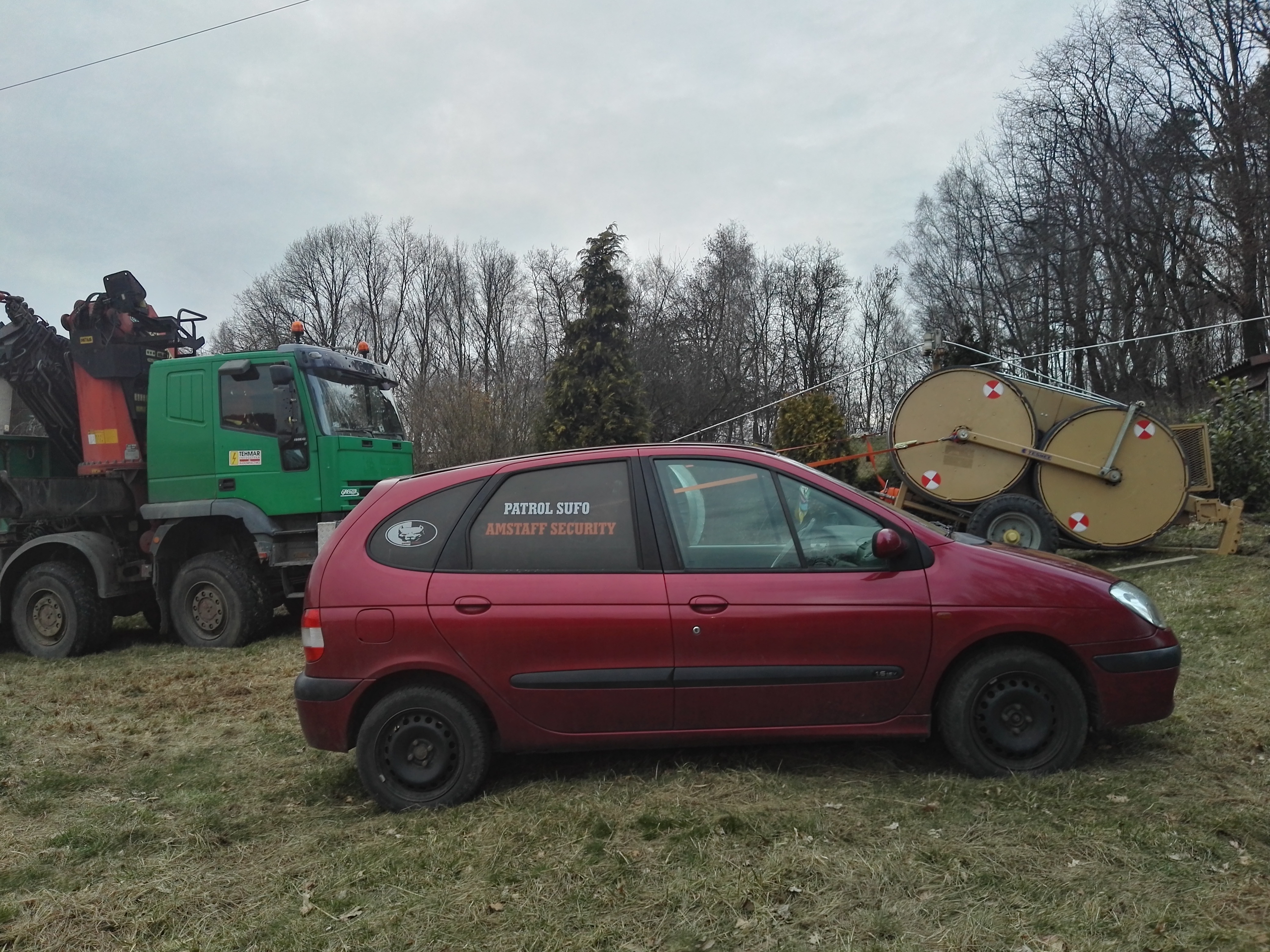Czerwony samochód osobowy z napisem 'PATROL SUFO AMSTAFF SECURITY' na szybie, zaparkowany obok zielonej ciężarówki z dźwigiem i przyczepy z dwoma dużymi bębnami na tle drzew.