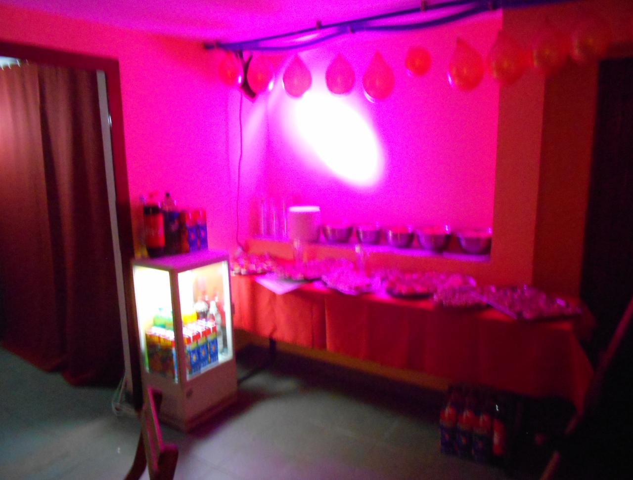 Sala oświetlona na różowo z dekoracjami balonowymi, bufetem z przekąskami, lodówką z napojami i zasłoniętym wejściem.