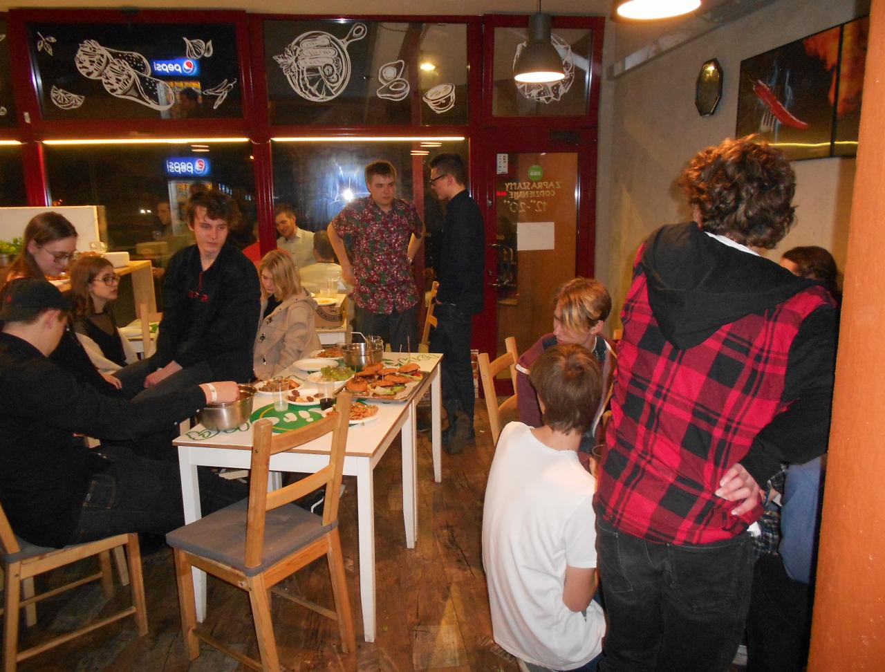 Grupa młodych ludzi siedząca przy stole z jedzeniem w lokalu, widoczne dekoracje na oknach i oświetlenie sufitowe.