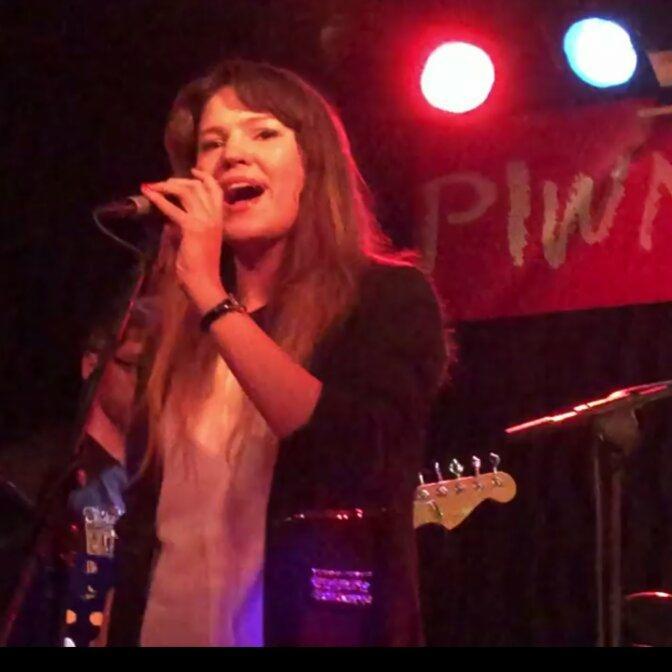 Wokalistka z mikrofonem na scenie podczas występu na żywo, w tle widoczny instrument i czerwone oświetlenie sceniczne.