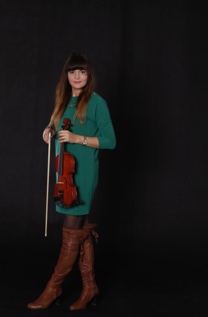 Kobieta w zielonej sukience i brązowych kozakach trzyma skrzypce i smyczek na czarnym tle.
