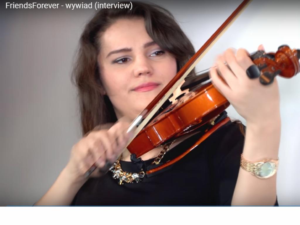 Kobieta gra na skrzypcach, ubrana w czarną bluzkę i ozdobny naszyjnik, z zegarkiem na ręce, w trakcie występu.