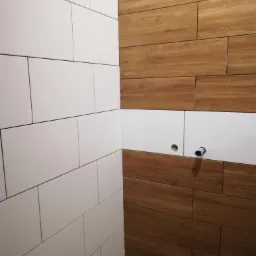Ściana łazienki wyłożona białymi, prostokątnymi płytkami ceramicznymi zestawionymi z płytkami drewnopodobnymi w ciepłym odcieniu brązu, widoczne przygotowane punkty instalacji wodnej.