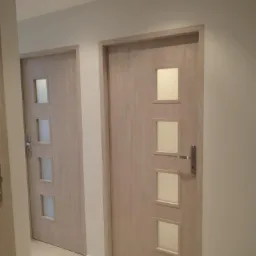 Dwa drewnopodobne drzwi wewnętrzne z czterema kwadratowymi szybkami, zamontowane w jasnym korytarzu z kremową podłogą i białymi ścianami.