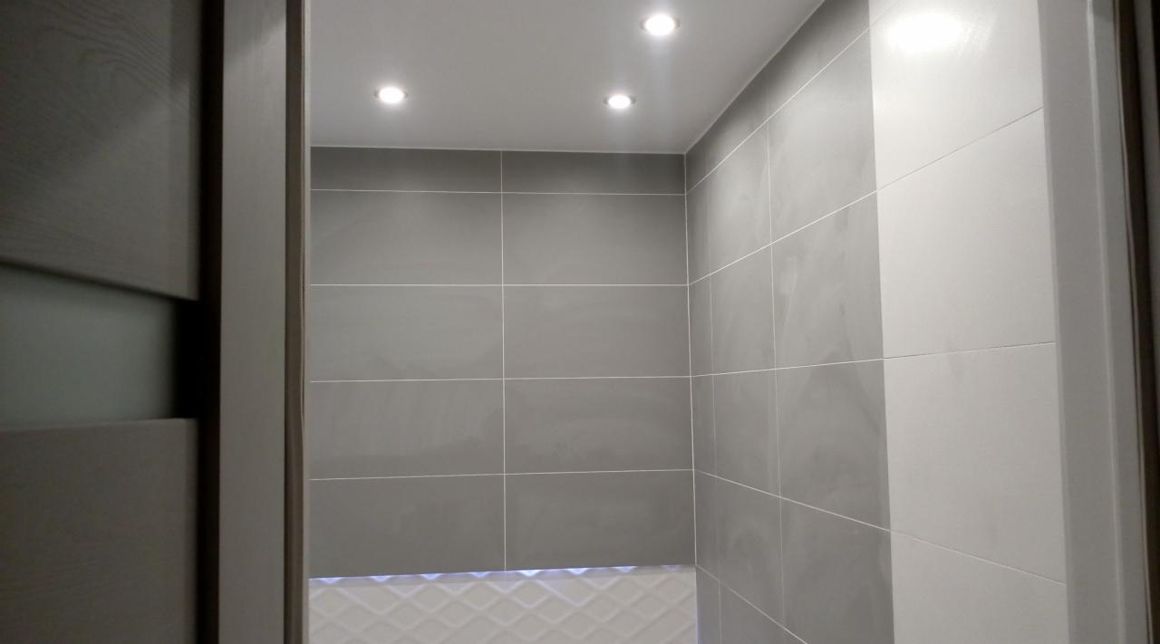 Wykończona łazienka z szarymi, kwadratowymi płytkami ułożonymi w regularną siatkę, oświetleniem punktowym w suficie i fragmentem drewnianych drzwi z widoczną fakturą.