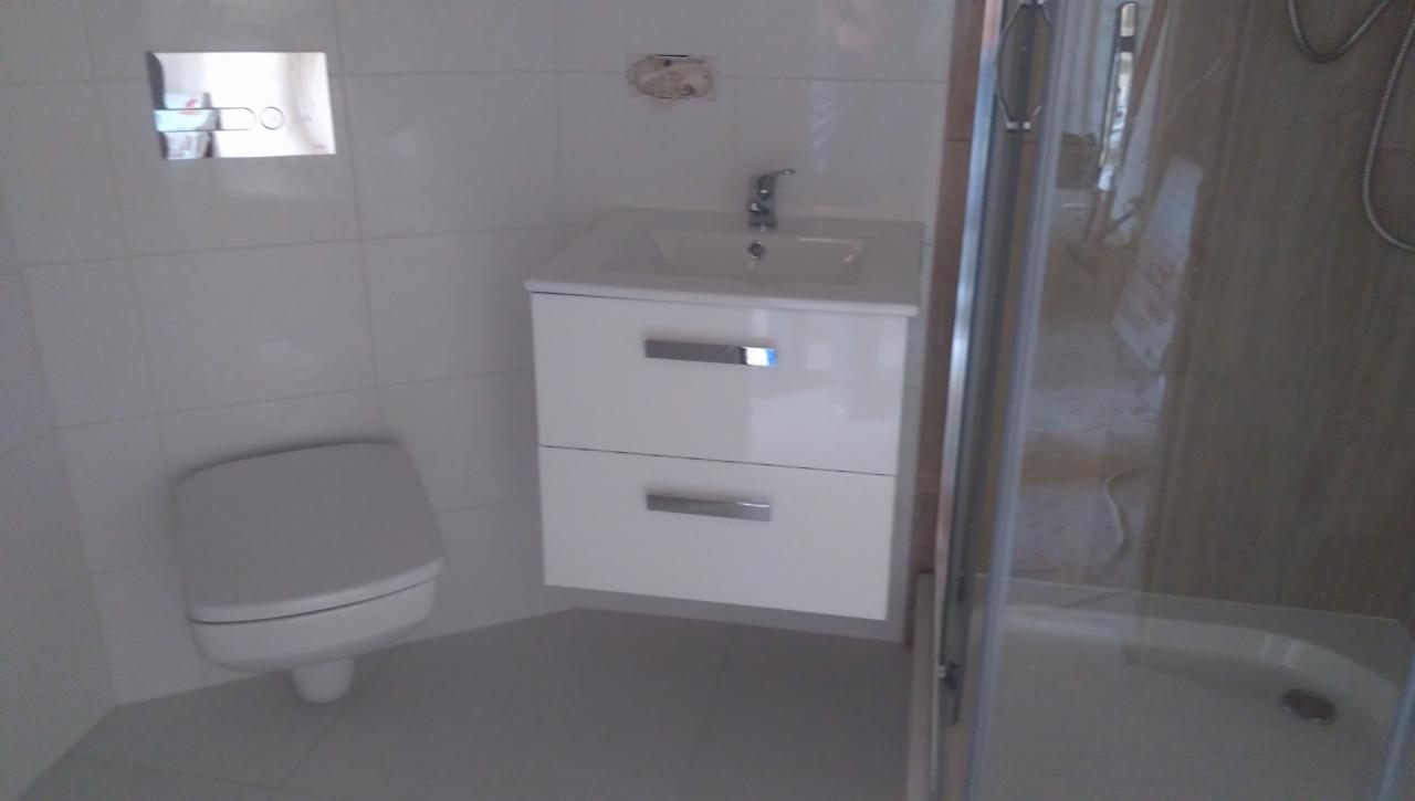 Nowoczesna łazienka z białą umywalką z szafką, podwieszanym WC i kabiną prysznicową z widoczną słuchawką.