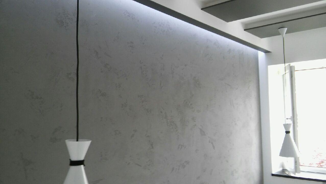 Ściana z szarą, dekoracyjną fakturą, oświetlona listwą LED pod sufitem, z wiszącą lampą w kształcie klepsydry i widocznym fragmentem okna.