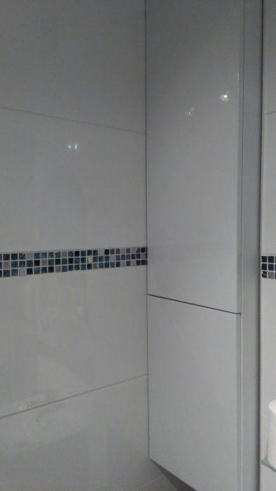 Biała szafka łazienkowa zamontowana przy ścianie z białymi płytkami i dekoracyjnym pasem mozaiki w odcieniach szarości i błękitu.