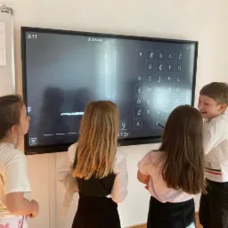 Dzieci w klasie, uczące się języka obcego przy interaktywnej tablicy z alfabetem i zadaniem do rozwiązania.