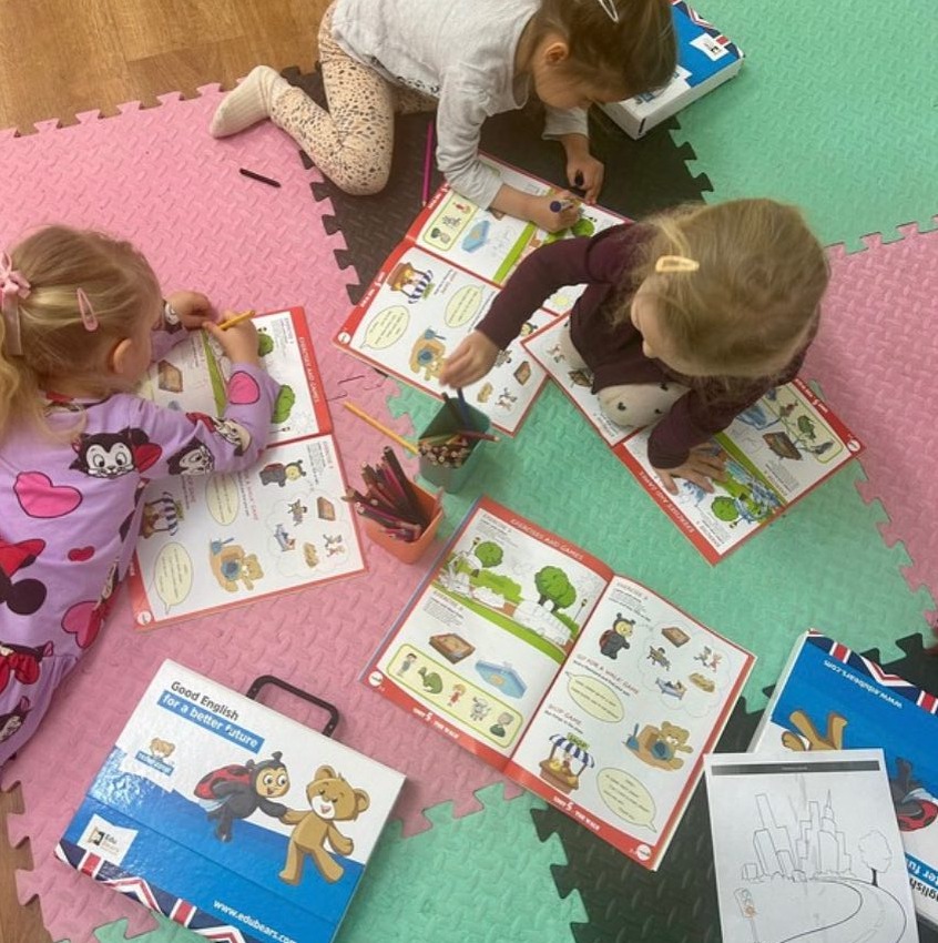 Trzy dziewczynki kolorują obrazki w książkach do nauki języka angielskiego na kolorowej macie podłogowej, widoczne kredki i materiały edukacyjne z napisem 'Good English for a better future'.