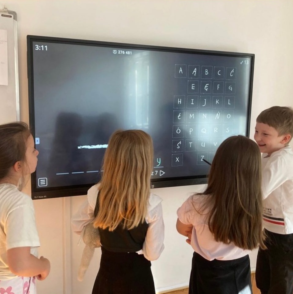 Dzieci w klasie, uczące się języka obcego przy interaktywnej tablicy z alfabetem i zadaniem do rozwiązania.